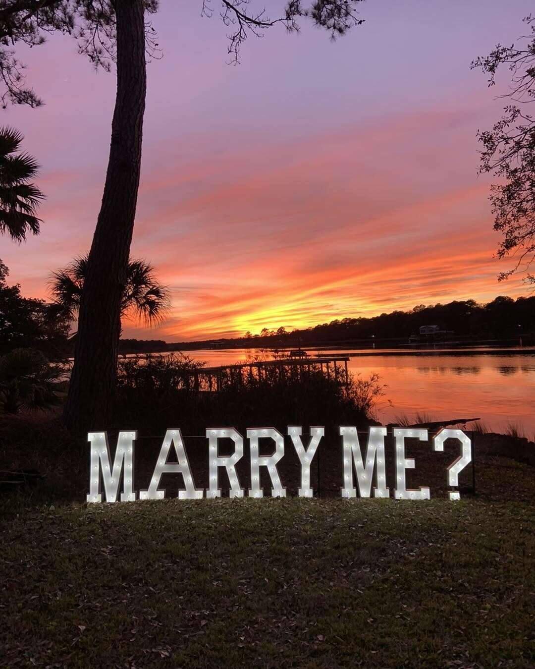 Lit marquee letters spelling MARRY ME on a lawn above the water at sunset
